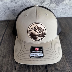 Può includere: Un cappello da camionista grigio e nero con una toppa in pelle marrone che rappresenta una scena di montagna. La toppa ha il testo "EST. 1970 RICHARDSON. ORIGINAL 112 TRUCKER ADJUSTABLE".