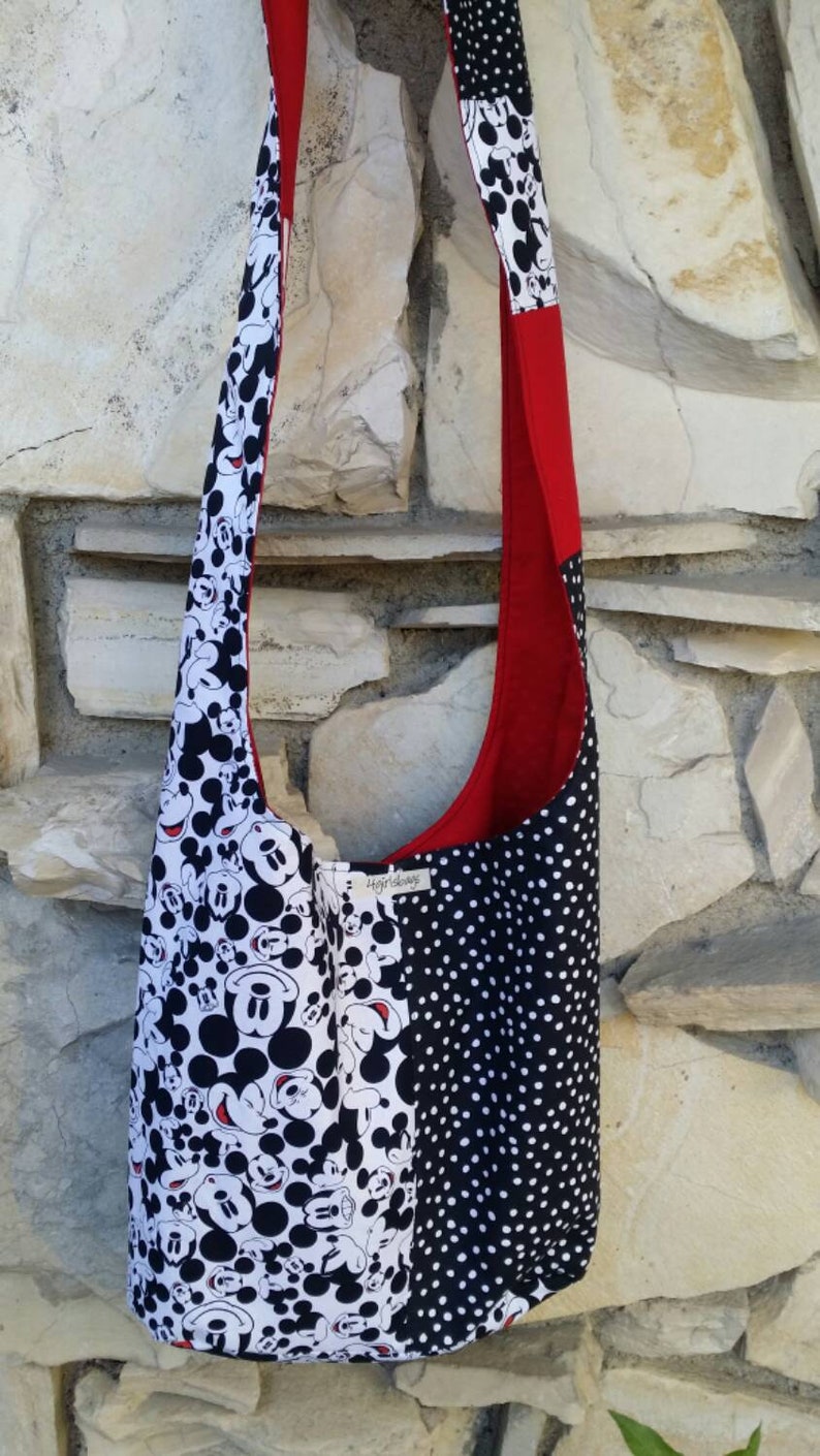 May include: A shoulder bag with a red interior and strap. The bag's exterior features a black and white Mickey Mouse print on one side and a black and white polka dot pattern on the other. The bag is made of fabric.