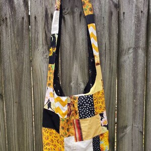 May include: A patchwork shoulder bag with a variety of fabrics, including sunflowers, polka dots, and chevron patterns. The bag features a long strap and a mix of yellow, black, and white colors. The bag is hanging on a wooden fence.