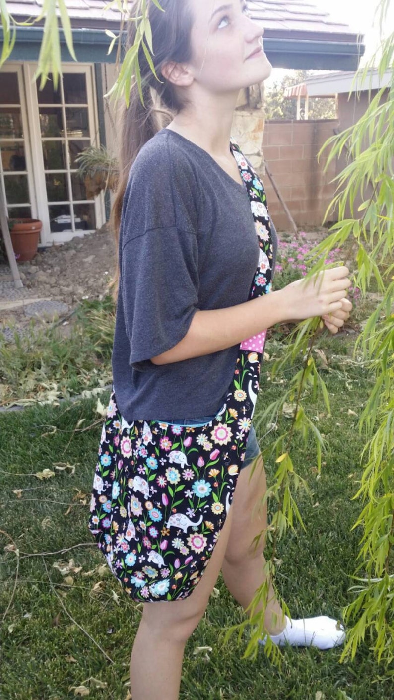 May include: A black tote bag with a colorful floral and elephant pattern. The bag has a wide strap with a pink polka dot design. The bag is worn over the shoulder.