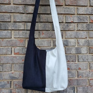 May include: A two-tone shoulder bag, split vertically with a black panel on the left and a white panel on the right. The bag hangs from a black strap. The background is a weathered brick wall.