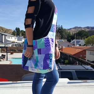 以下が含まれることがあります： 黒いカットアウトスリーブのシャツと青いジーンズを着た女性が、紫、青、白の生地で作られたパッチワークのトートバッグを持っています。バッグは長いストラップが付いており、肩にかけられています。