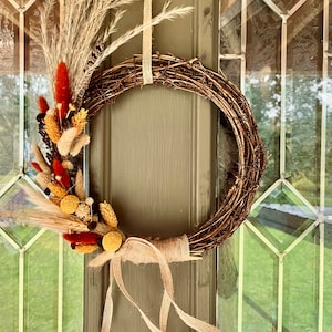 May include: A rustic grapevine wreath adorned with dried flowers and wheat stalks in shades of orange, beige, and yellow. The wreath is tied with a burlap ribbon and hangs on a green door with glass panels, creating a warm, natural aesthetic.