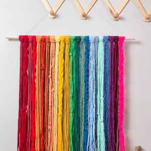 May include: A rainbow-colored macrame wall hanging. The wall decor features a wooden dowel with multiple strands of yarn in various colors, including red, orange, yellow, green, blue, and purple. Some strands are braided, and others are knotted.