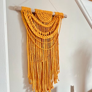 May include: A mustard yellow macrame wall hanging with a wooden dowel at the top and long fringe at the bottom.
