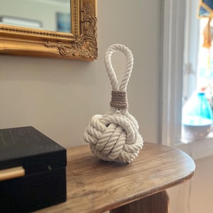 May include: A decorative white rope knot doorstop with a loop handle, wrapped with brown twine. The nautical-themed doorstop sits on a wooden table, next to a black box with a gold handle.