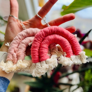 May include: Two macrame rainbow keychains in shades of pink and coral, with cream-colored fringe. The keychains are held up against a blurred background of greenery and a window, showcasing the handmade craft and vibrant colors.