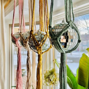 May include: Four macrame plant hangers with disco ball ornaments. The hangers are made with pink, cream, yellow, and green cord. The disco balls are silver and reflect light.