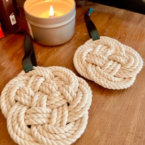May include: Two off-white rope coasters with a decorative knot design and dark green ribbon loops. A lit candle in a silver tin sits nearby. The coasters are on a wooden surface.