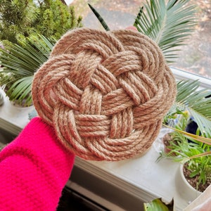 May include: A close-up of a round, woven coaster made from natural jute rope. The coaster has a knot pattern and is being held by a hand wearing a pink sweater.