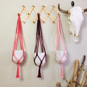May include: Three macrame plant hangers with white ceramic pots. The hangers are pink, burgundy, and light pink. The hangers are hanging from a wooden rack on a white wall.
