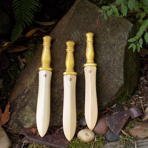 Puede incluir: Tres espadas de madera con hojas de color beige claro y empuñaduras pintadas de dorado. Cada espada presenta un diseño decorativo cerca de la hoja. Las espadas se exhiben al aire libre contra una gran roca y vegetación.