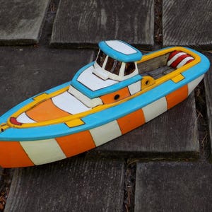 Könnte beinhalten: Ein dekoratives Modellboot mit einem Rumpf, der mit abwechselnden orangefarbenen und weißen vertikalen Streifen bemalt ist. Das Deck ist mit hellblauen Verzierungen, einer weißen Kabine mit dunklen Fenstern und einem rot-weiß gestreiften Kissen am Heck versehen.