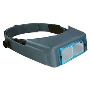 May include: A gray headband magnifier with two clear lenses. The headband is adjustable and has a black strap.