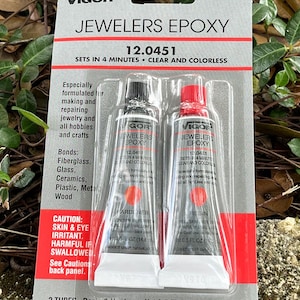 May include: Two tubes of clear epoxy adhesive, labeled "Jewelers Epoxy" and "12.0451". The packaging states that the epoxy sets in 4 minutes and is clear and colorless. It is formulated for making and repairing jewelry and crafts. The tubes are labeled "Resin" and "Hardener" and contain 0.5 fluid ounces (14.8 milliliters) each.