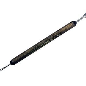 May include: A Kemper Tools USA K27 dual-ended sculpting tool. One end features a pointed, spear-like tip, while the other has a thin, wire-like tip. The handle is dark wood-toned with the brand name and model number printed on it.