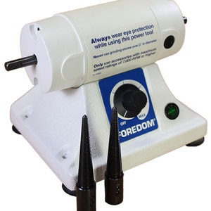 Może przedstawiać: Elektryczna szlifierka stołowa w kolorze białym i niebieskim z czarnym pokrętłem i zielonym światełkiem. Szlifierka ma dwa czarne trzpienie i naklejkę ostrzegawczą z napisem "Zawsze noś ochronę oczu podczas używania tego narzędzia elektrycznego. Nigdy nie używaj kamieni szlifierskich o średnicy większej niż 5 cm. Używaj tylko akcesori&oacute;w o maksymalnej prędkości 7 000 obr./min lub wyższej."