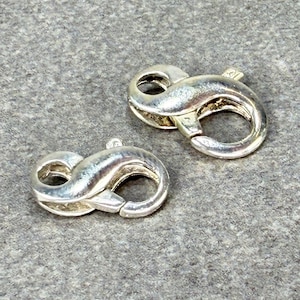 May include: Two silver-colored lobster claw clasps. These clasps are in the shape of an infinity symbol. The clasps are made of a shiny metal and are designed to connect jewelry pieces. The background is a textured gray surface.