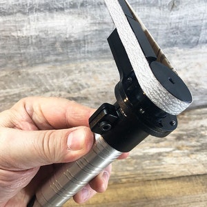 May include: A black and silver handheld belt sander with a gray sanding belt. The sander has a textured grip and a trigger.