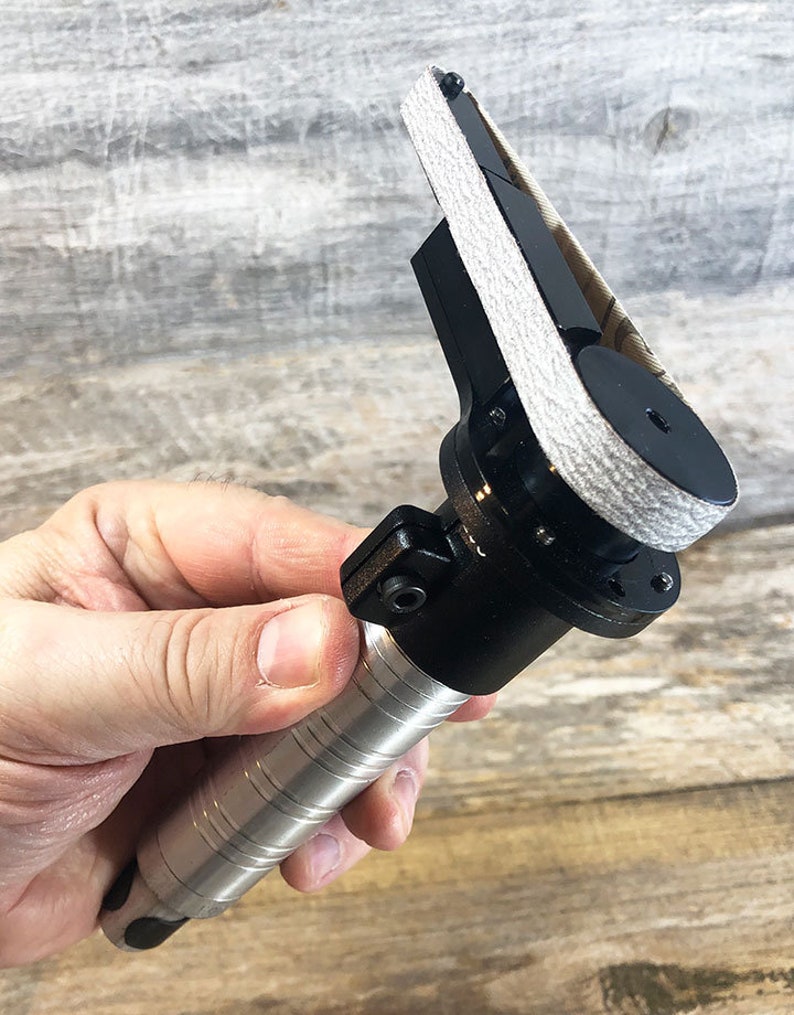 May include: A black and silver handheld belt sander with a gray sanding belt. The sander has a textured grip and a trigger.