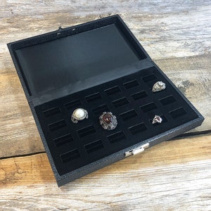 May include: Black ring box with a velvet interior and multiple compartments for storing rings. The box has a silver latch and is open to reveal several rings with different gemstones.