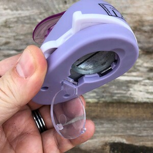 Pode incluir: Um perfurador de papel artesanal lavanda e branco com um mecanismo de corte circular de metal e uma porta de pl&aacute;stico transparente na parte inferior. O dispositivo tem uma alavanca roxa no topo e texto impresso na lateral da carca&ccedil;a.