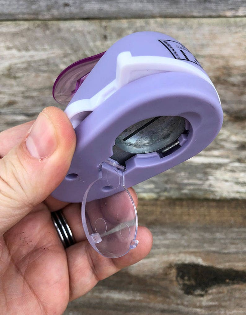 Pode incluir: Um perfurador de papel artesanal lavanda e branco com um mecanismo de corte circular de metal e uma porta de pl&aacute;stico transparente na parte inferior. O dispositivo tem uma alavanca roxa no topo e texto impresso na lateral da carca&ccedil;a.