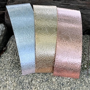 May include: Three metal rectangles with embossed floral patterns. The rectangles are silver, gold, and copper-colored. The silver rectangle is on the left, the gold in the middle, and the copper on the right. The rectangles are displayed on a bed of small rocks.