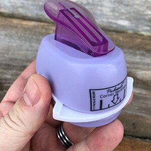 Pode incluir: Um perfurador de papel roxo claro com uma al&ccedil;a superior roxa transl&uacute;cida e uma base branca. A lateral apresenta uma etiqueta com os dizeres Packasmile Corner Necklace e U.S. Pat. No. 8,763,503 com um &iacute;cone de seta apontando para baixo.