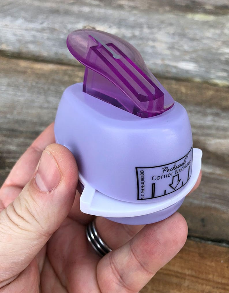Pode incluir: Um perfurador de papel roxo claro com uma al&ccedil;a superior roxa transl&uacute;cida e uma base branca. A lateral apresenta uma etiqueta com os dizeres Packasmile Corner Necklace e U.S. Pat. No. 8,763,503 com um &iacute;cone de seta apontando para baixo.