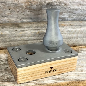 May include: A silver metal fret press with a wooden base. The base is labeled "Fretz" and the press is labeled "SRM 1".