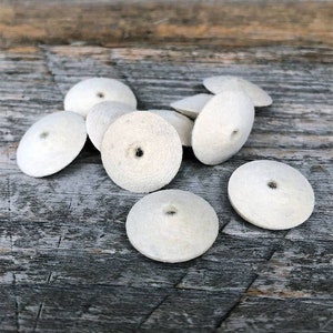 May include: A group of eight white felt polishing wheels, each with a small hole in the center. The wheels are arranged in a loose pile on a weathered wooden surface.