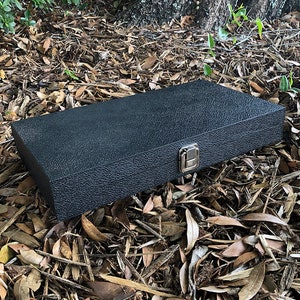 K&ouml;nnte beinhalten: Eine schwarze rechteckige Box mit einer strukturierten Oberfl&auml;che und einem silbernen Verschluss. Die Box liegt auf einem Bett aus braunen Bl&auml;ttern.