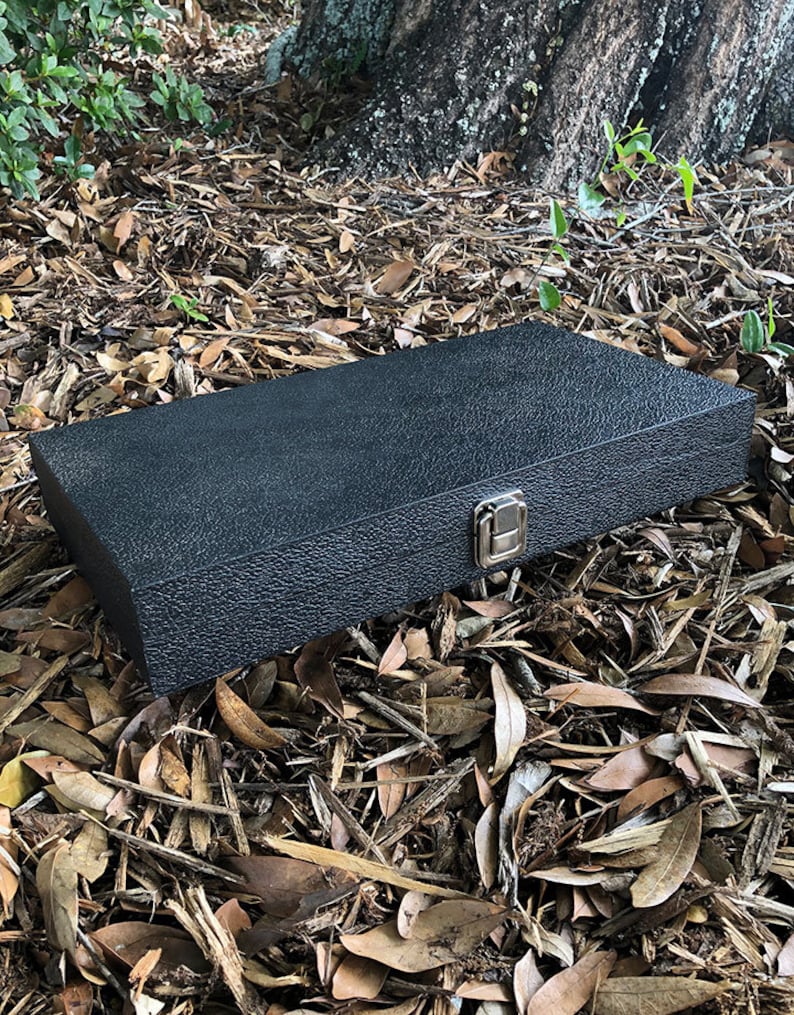 K&ouml;nnte beinhalten: Eine schwarze rechteckige Box mit einer strukturierten Oberfl&auml;che und einem silbernen Verschluss. Die Box liegt auf einem Bett aus braunen Bl&auml;ttern.