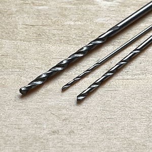 May include: Three drill bits of different sizes, lying on a light wood surface. The drill bits are made of metal and have a silver color.