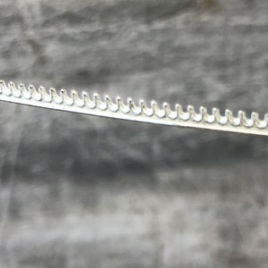 Puede incluir: Primer plano de una tira de metal plateado con una fila de pequeños dientes espaciados uniformemente a lo largo del borde.