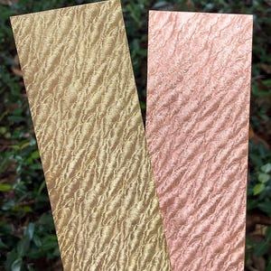 May include: Two rectangular wood pieces with a wavy grain pattern. One is a light gold color, and the other is a soft pink. The wood grain has a textured appearance. The background is blurred green foliage.