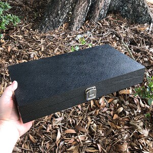 K&ouml;nnte beinhalten: Eine schwarze rechteckige Box mit einer strukturierten Oberfl&auml;che und einem goldenen Verschluss. Die Box ist aus robustem Material gefertigt und hat ein elegantes, modernes Design.
