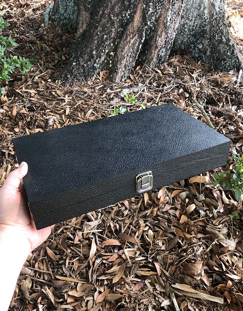 K&ouml;nnte beinhalten: Eine schwarze rechteckige Box mit einer strukturierten Oberfl&auml;che und einem goldenen Verschluss. Die Box ist aus robustem Material gefertigt und hat ein elegantes, modernes Design.
