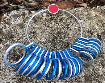 May include: A silver ring sizer with a red top and blue and silver rings. The sizer is on a gray stone surface.