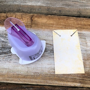 Pode incluir: Uma ferramenta de perfura&ccedil;&atilde;o artesanal em violeta claro e branco com uma pega superior violeta transl&uacute;cida, rotulada como Pocket Necklace Corner Punch, ao lado de uma etiqueta de papel retangular creme com duas pequenas ranhuras.