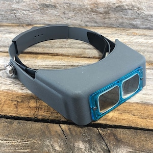 May include: Gray headband magnifier with two blue magnifying lenses. The headband is adjustable and has a black strap.