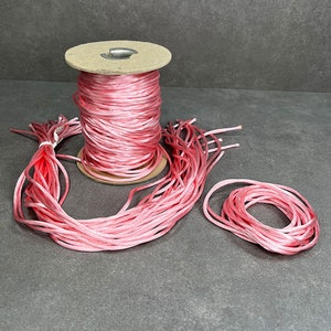 May include: A spool of pink satin cord, with some of the cord unwound and lying on a gray surface.