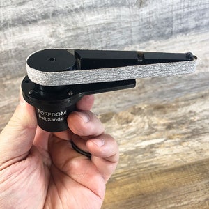May include: A black Foredom belt sander with a gray sanding belt. The sander is small and handheld.