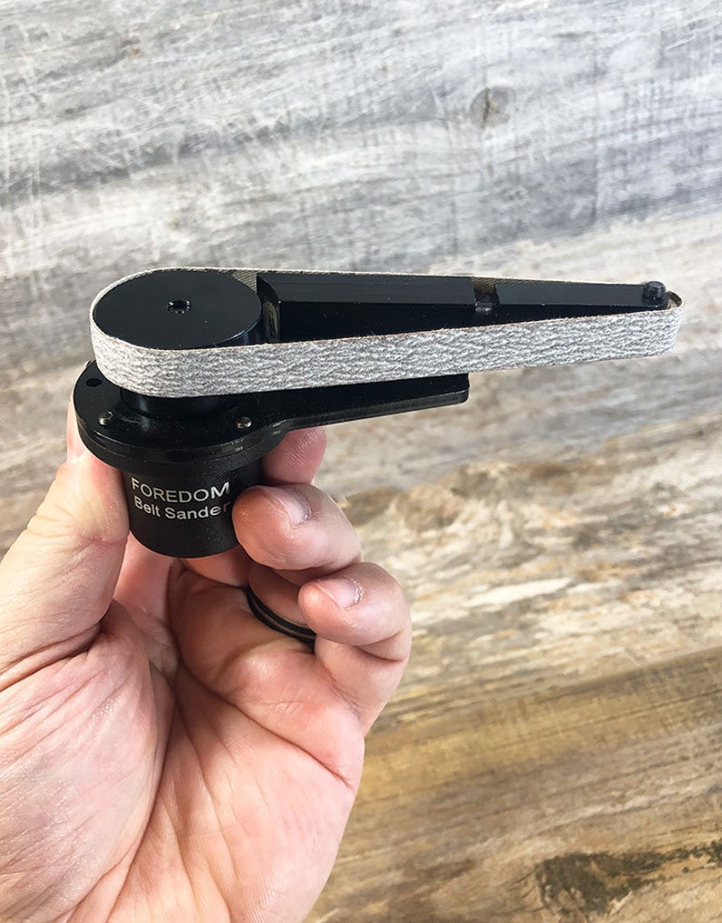 May include: A black Foredom belt sander with a gray sanding belt. The sander is small and handheld.