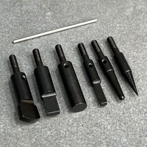 May include: A set of seven black metal tools with different shaped tips. The tools are arranged in a row on a gray surface. The tools are used for leather crafting.