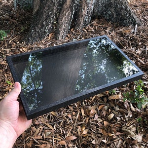 May include: A black rectangular display case with a clear glass top. The case is made of a textured material and has a sleek, modern design.