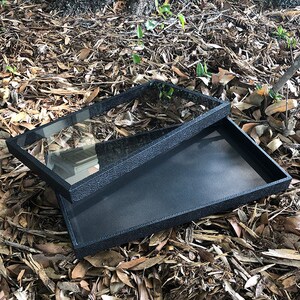 May include: Two black trays with a textured finish. The top tray has a clear glass insert. The trays are on a bed of brown leaves.