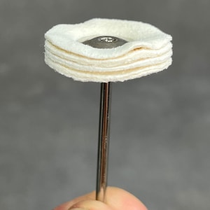May include: A close-up of a white felt buffing wheel attached to a metal rod. The buffing wheel is made of multiple layers of felt.