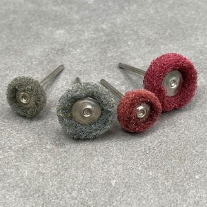 May include: Four abrasive buffing wheels with metal stems. The wheels are in various sizes and colors, including gray and red. The wheels have a circular shape and a textured surface, designed for polishing or cleaning.